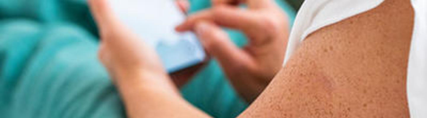 Člověk ležící na pohovce se zvednutými nohami, na paži má senzor pro monitorování glukózy a používá chytrý telefon.